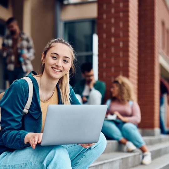 Happy female student surfing the net on laptop in front of university building and looking at camera.